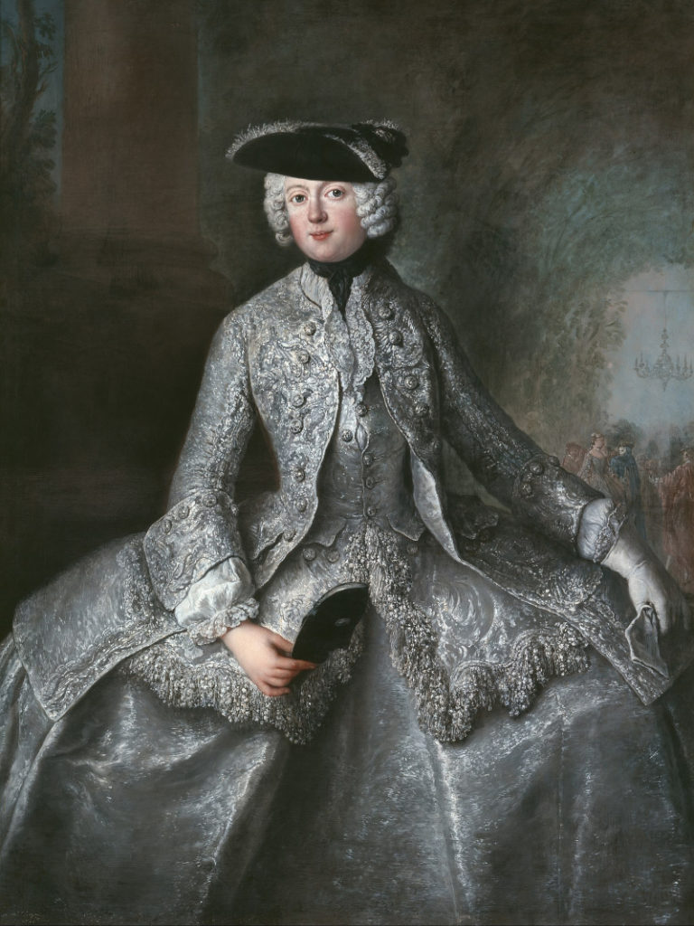 Portrait of Anna Amalia of Prussia as a young woman. She looks directly at the viewer, and wears a powdered wig and black tricorn hat. Her gray, longsleeve, high-necked dress is heavily embroidered and her skirt spreads out wide at the hips.
